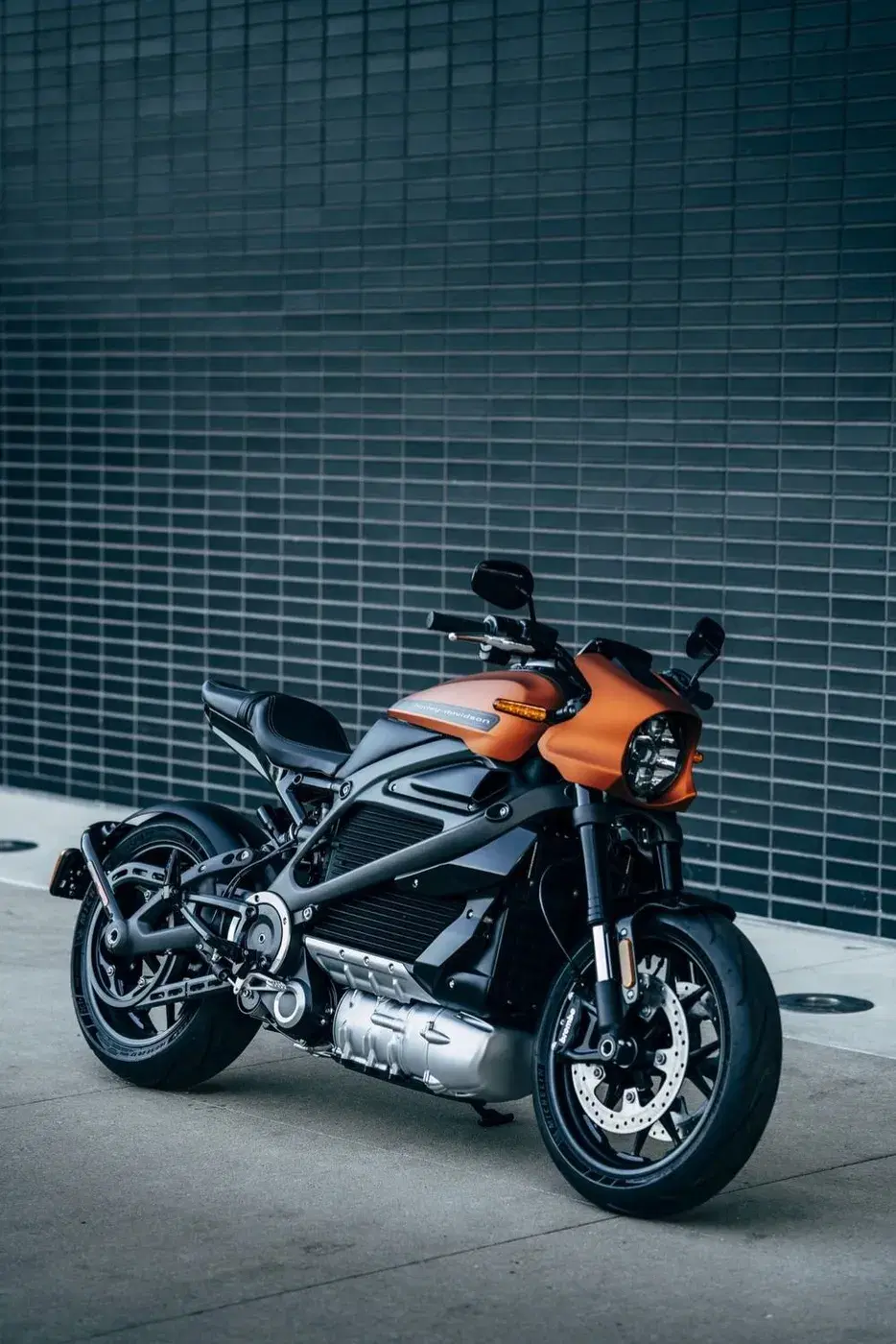 An orange and black electric motorcycle parked against a tiled wall
